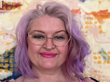 A person with pastel purple hair, cat-eye glasses, and a nose ring smiles in front of a colorful abstract background. They wear a striped top with thin straps and have a visible tattoo on their right arm.