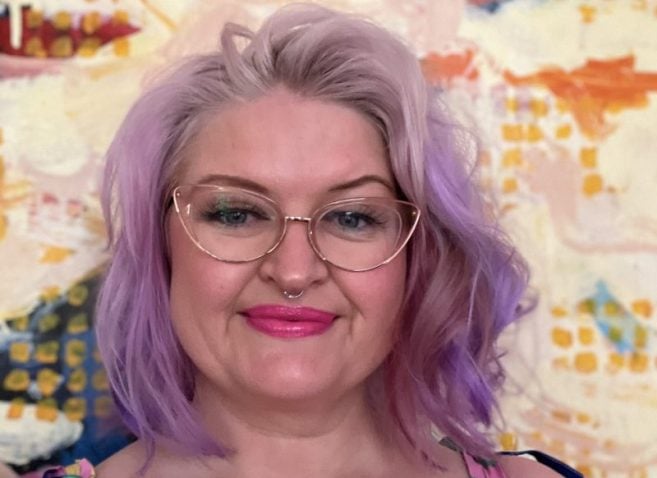 A person with pastel purple hair, cat-eye glasses, and a nose ring smiles in front of a colorful abstract background. They wear a striped top with thin straps and have a visible tattoo on their right arm.