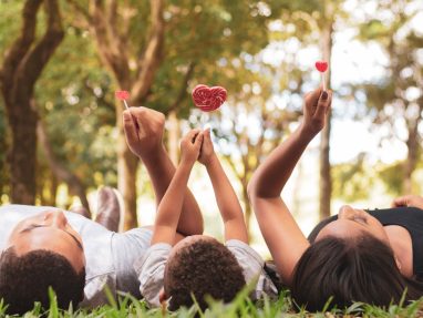 Three people lying on grass in a park, holding up red heart-shaped lollipops. Trees and sunlight are visible in the background, creating a warm and joyful atmosphere.