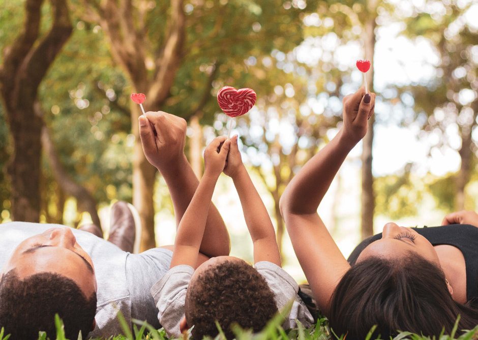 Three people lying on grass in a park, holding up red heart-shaped lollipops. Trees and sunlight are visible in the background, creating a warm and joyful atmosphere.