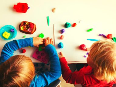 Two young children sit at a table playing with colorful modeling clay, shaping and arranging pieces on the tabletop and a white tray, surrounded by various clay tools and small containers.