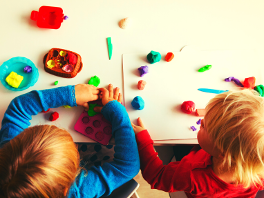 Two young children sit at a table playing with colorful modeling clay, shaping and arranging pieces on the tabletop and a white tray, surrounded by various clay tools and small containers.