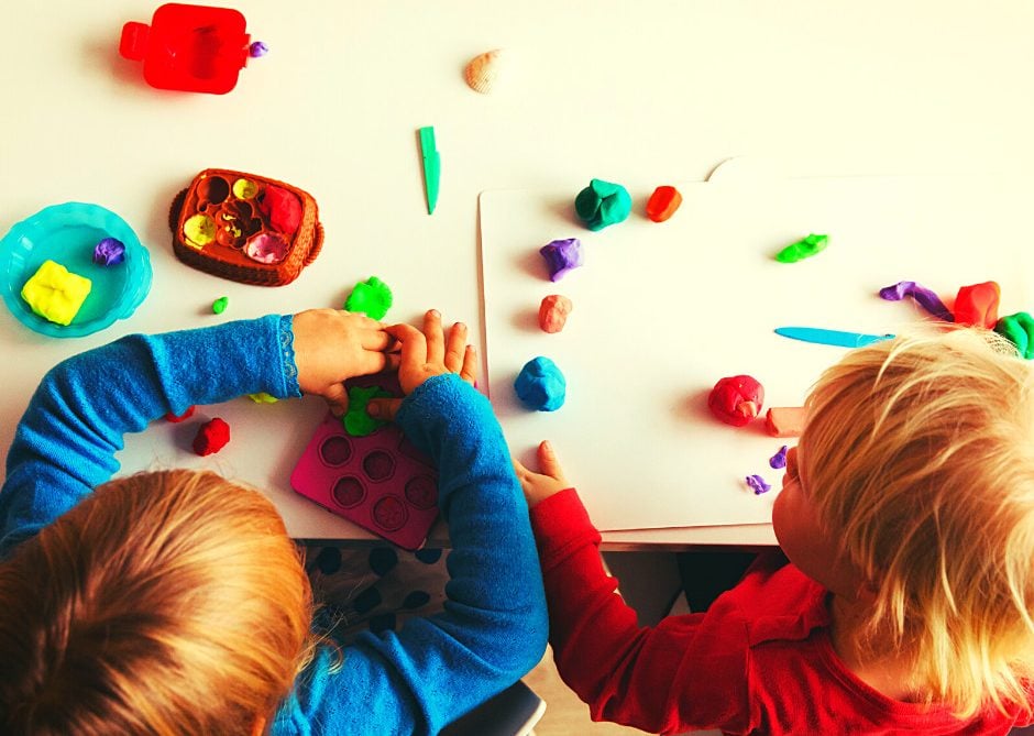 Two young children sit at a table playing with colorful modeling clay, shaping and arranging pieces on the tabletop and a white tray, surrounded by various clay tools and small containers.