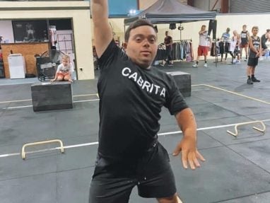 A man wearing a black CABRITA shirt lifts a dumbbell overhead in a gym, surrounded by workout equipment and people in the background.
