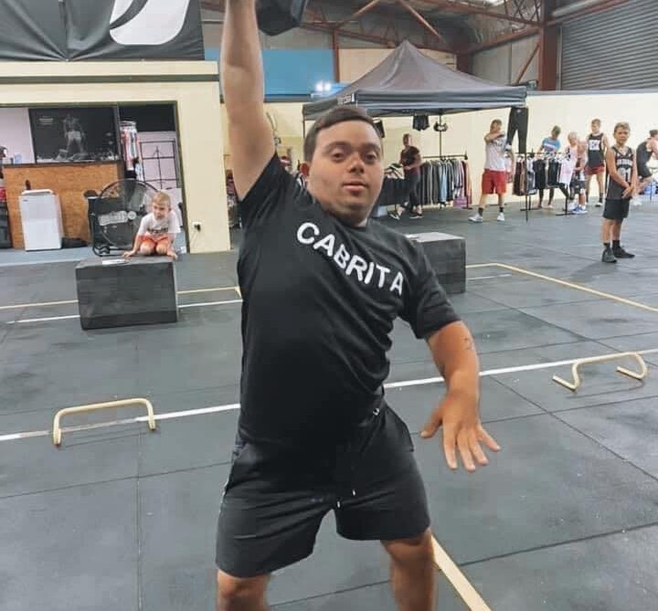 A man wearing a black CABRITA shirt lifts a dumbbell overhead in a gym, surrounded by workout equipment and people in the background.