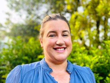 A woman with light skin and brown hair pulled back smiles at the camera, wearing a blue blouse, standing outdoors in front of lush green foliage and trees.