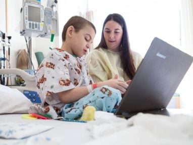 A young child in hospital pajamas sits on a hospital bed using a laptop, accompanied by a woman in a gown. Medical equipment is visible in the background, suggesting a hospital setting.