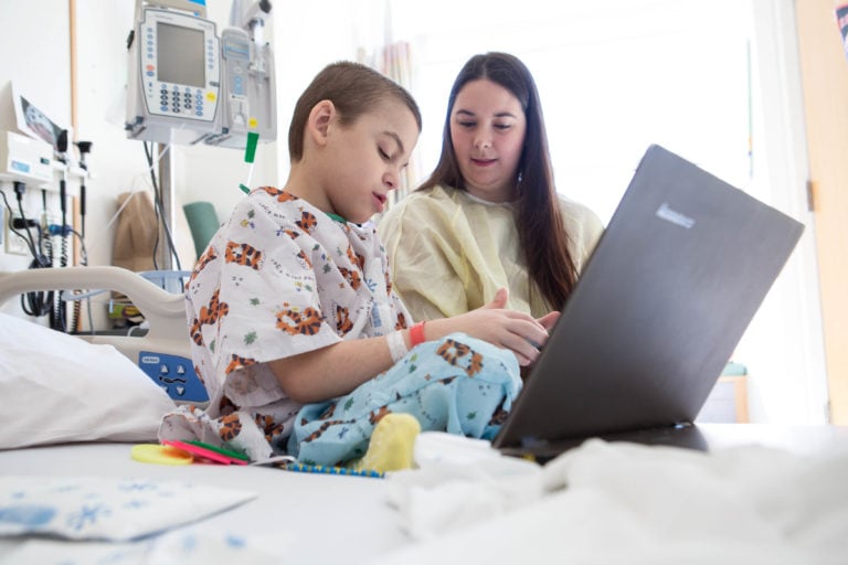 A young child in hospital pajamas sits on a hospital bed using a laptop, accompanied by a woman in a gown. Medical equipment is visible in the background, suggesting a hospital setting.