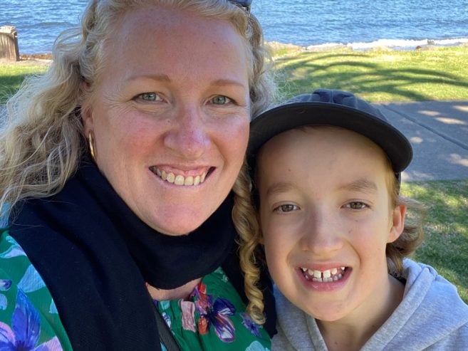 A smiling woman with curly blonde hair and a boy with a missing front tooth stand close together outdoors near water, grass, and a sidewalk.