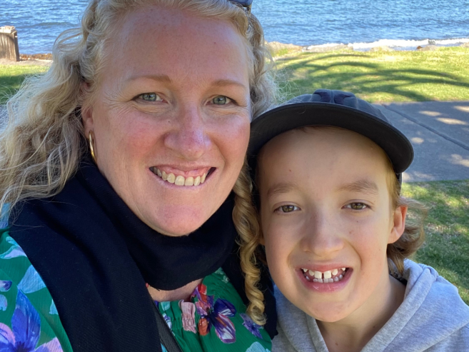 A smiling woman with curly blonde hair and a boy with a missing front tooth stand close together outdoors near water, grass, and a sidewalk.