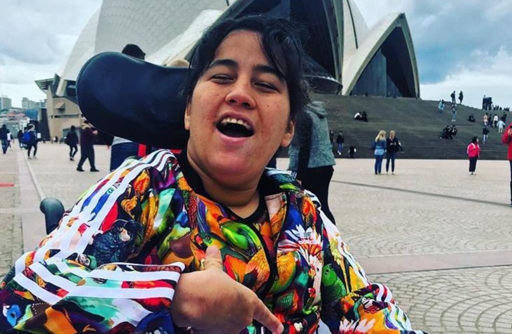 A smiling woman in a colorful jacket sits in a wheelchair, pointing at herself, with the Sydney Opera House and people walking in the background on a cloudy day.