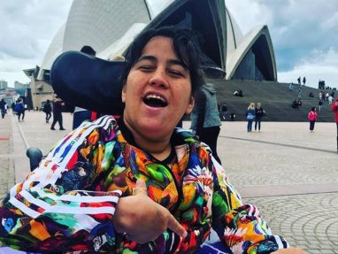 A smiling woman in a colorful jacket sits in a wheelchair, pointing at herself, with the Sydney Opera House and people walking in the background on a cloudy day.