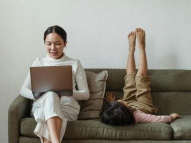 A woman sits on a couch working on a laptop while a child lies beside her with legs up against the couch backrest, creating a relaxed and playful atmosphere at home.