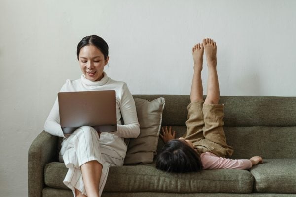A woman sits on a couch working on a laptop while a child lies beside her with legs up against the couch backrest, creating a relaxed and playful atmosphere at home.