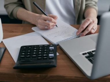 A person writes notes on paper at a desk with a laptop, calculator, and documents, suggesting they are working on financial or accounting tasks.