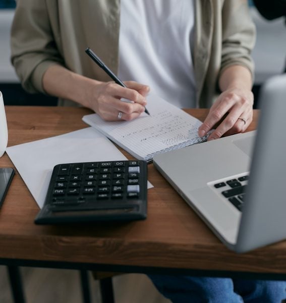 A person writes notes on paper at a desk with a laptop, calculator, and documents, suggesting they are working on financial or accounting tasks.