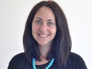 A woman with straight, shoulder-length dark hair smiles at the camera. She is wearing a black top, a black cardigan, and a chunky turquoise beaded necklace, standing against a plain white background.