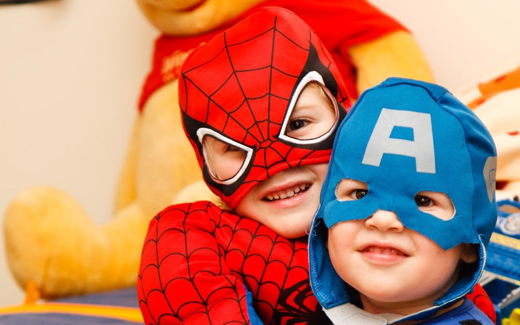 Two young children smile while wearing Spider-Man and Captain America costumes. A large yellow stuffed animal is visible in the background.