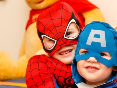 Two young children smile while wearing Spider-Man and Captain America costumes. A large yellow stuffed animal is visible in the background.