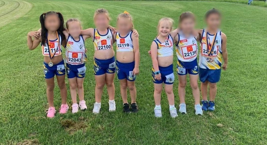 Six young children in athletic uniforms stand in a row on green grass, smiling and posing for a photo. Their faces are blurred, and each child is wearing a race number on their shorts or shirt.
