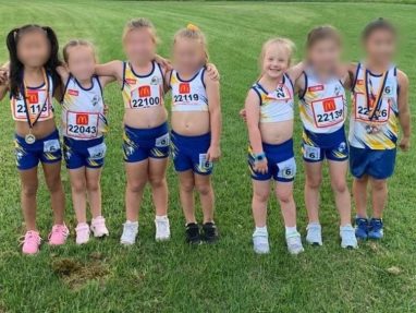 Six young children in athletic uniforms stand in a row on green grass, smiling and posing for a photo. Their faces are blurred, and each child is wearing a race number on their shorts or shirt.