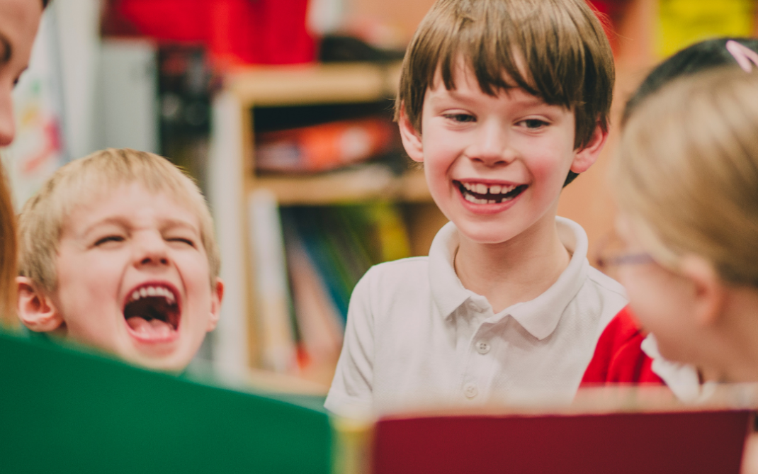 A group of young children in a classroom smile and laugh together while looking at a colorful book being held by an adult. Shelves with books and bins are visible in the background.