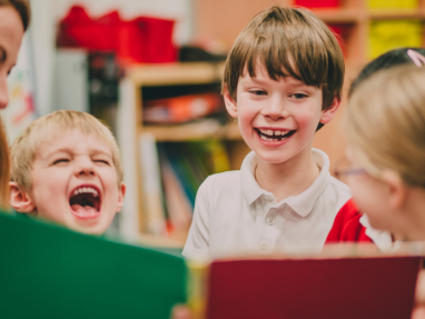 A group of young children in a classroom smile and laugh together while looking at a colorful book being held by an adult. Shelves with books and bins are visible in the background.