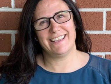 A woman with long dark hair and glasses smiles in front of a red brick wall, wearing a dark teal top.