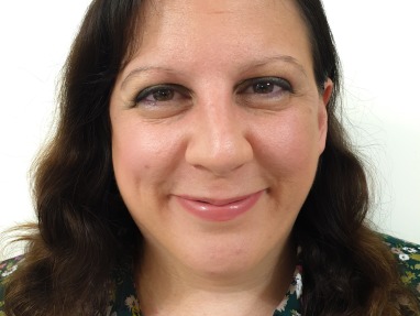 A woman with wavy brown hair smiles at the camera. She is wearing a green floral blouse and has natural makeup. The background is plain white.