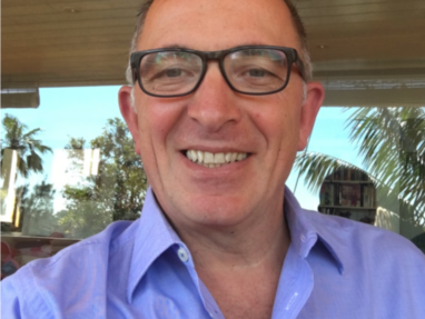 A man wearing glasses and a light purple collared shirt smiles at the camera. He is indoors with large windows behind him, showing palm trees and bookshelves in the background.