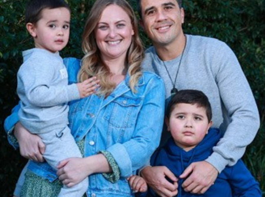 A smiling family of four poses outdoors in front of green bushes. The mother holds a young boy while standing next to the father, who holds an older boy’s shoulders. The word envisage with a jumping figure is overlaid on the image.