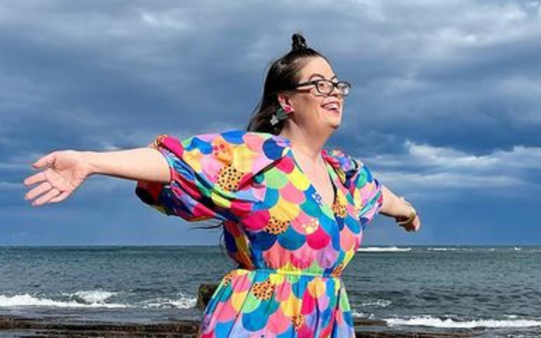 A smiling woman in glasses and a colorful dress stands on rocky ground near the ocean, arms outstretched, with a cloudy sky above.