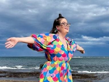A smiling woman in glasses and a colorful dress stands on rocky ground near the ocean, arms outstretched, with a cloudy sky above.