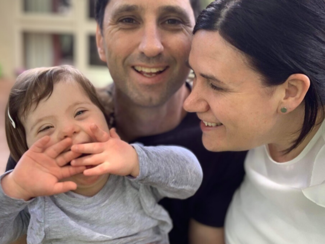 A smiling couple poses closely with a laughing young girl who has Down syndrome. The girl holds her hands up playfully in front of her face. They all appear happy and are enjoying a joyful moment together.