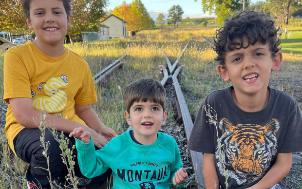 Three young boys sit and smile together on old, overgrown railroad tracks in a grassy area. The boy on the left wears a yellow shirt, the middle one wears a teal shirt, and the boy on the right wears a black tiger shirt.