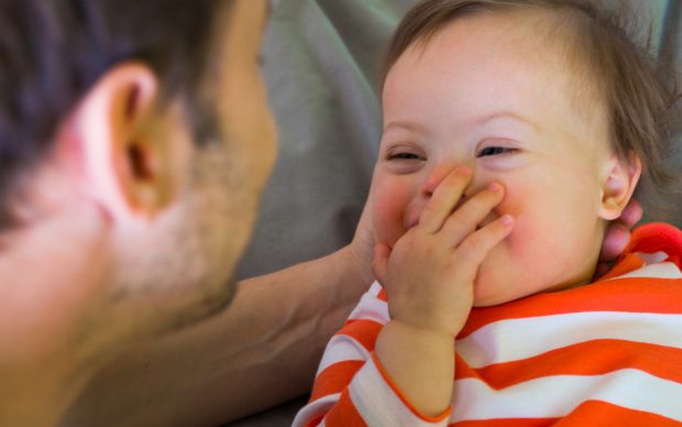 A smiling baby in an orange and white striped shirt covers their mouth with one hand while looking at an adult who is facing them closely. The interaction appears joyful and affectionate.