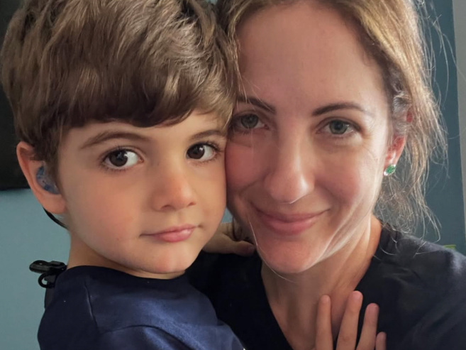A young child with brown hair and a hearing aid hugs a smiling woman with light brown hair and green earrings. They are close together, looking at the camera.