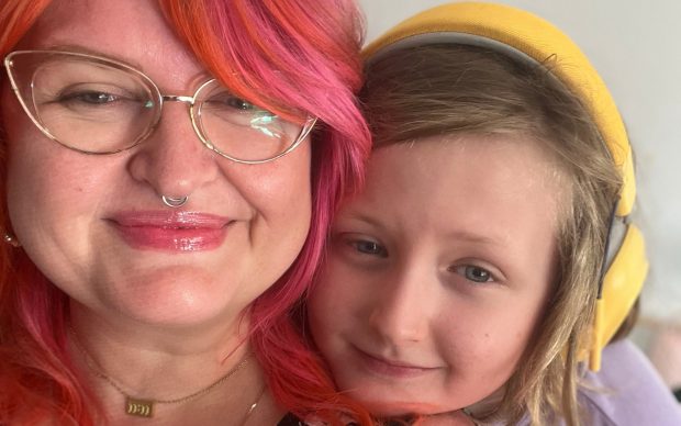 A smiling adult with bright pink and orange hair and glasses poses for a close-up selfie with a child wearing yellow headphones, both appearing happy and relaxed.