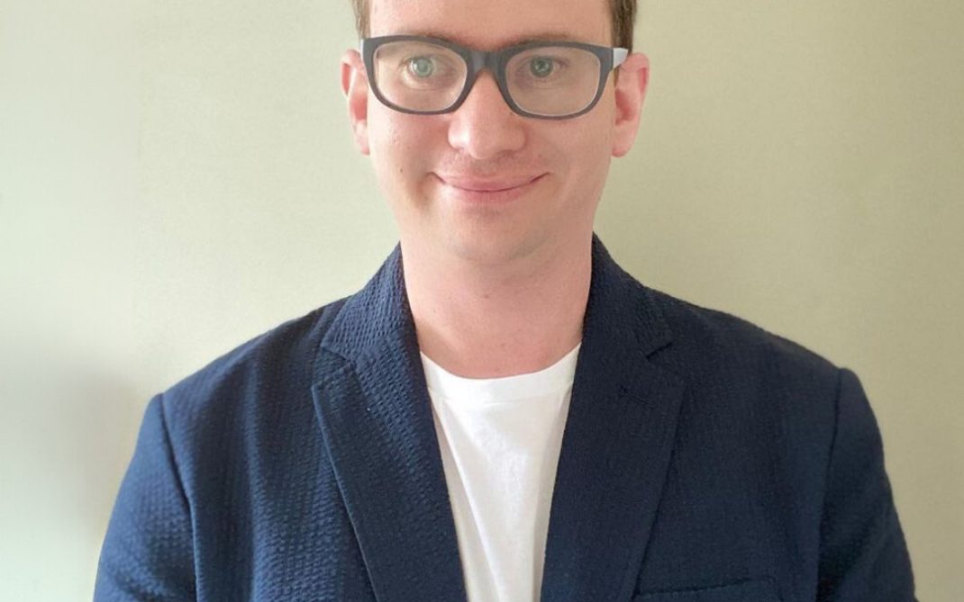 A person with short light brown hair and glasses is wearing a navy blue blazer over a white shirt, standing in front of a plain light-colored wall and smiling slightly.