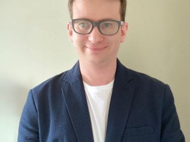 A person with short light brown hair and glasses is wearing a navy blue blazer over a white shirt, standing in front of a plain light-colored wall and smiling slightly.