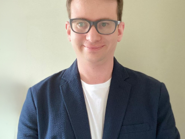 A person with short light brown hair and glasses is wearing a navy blue blazer over a white shirt, standing in front of a plain light-colored wall and smiling slightly.