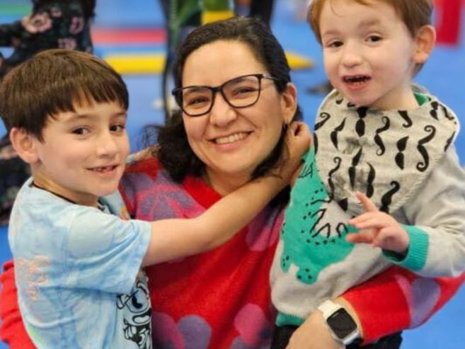 A woman wearing glasses and a red sweater smiles while hugging two young boys, one on each side, in a colorful indoor setting. The boys also smile at the camera.