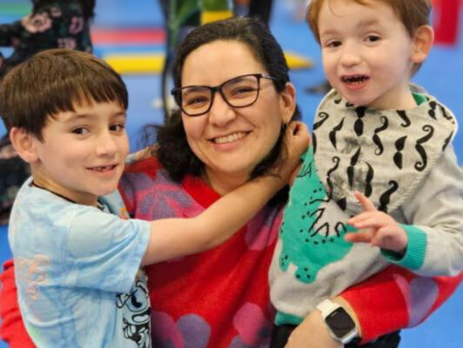 A woman wearing glasses and a red sweater smiles while hugging two young boys, one on each side, in a colorful indoor setting. The boys also smile at the camera.