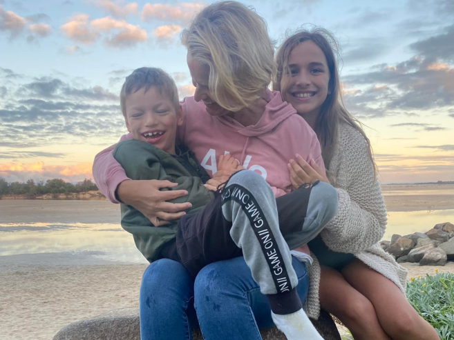 A woman sits on a rock at the beach, smiling and holding a laughing young boy on her lap, while an older girl hugs them from the side. The sky is cloudy and the beach is visible in the background.