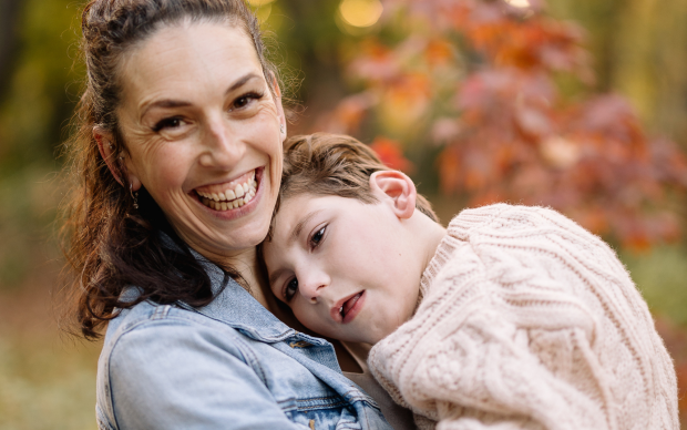 A woman smiles warmly while holding a child who rests their head on her shoulder. They are outdoors with autumn-colored leaves in the background.