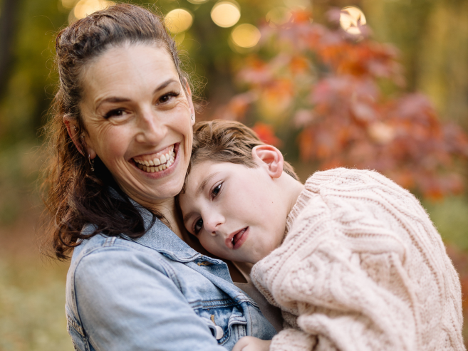 A woman smiles warmly while holding a child who rests their head on her shoulder. They are outdoors with autumn-colored leaves in the background.