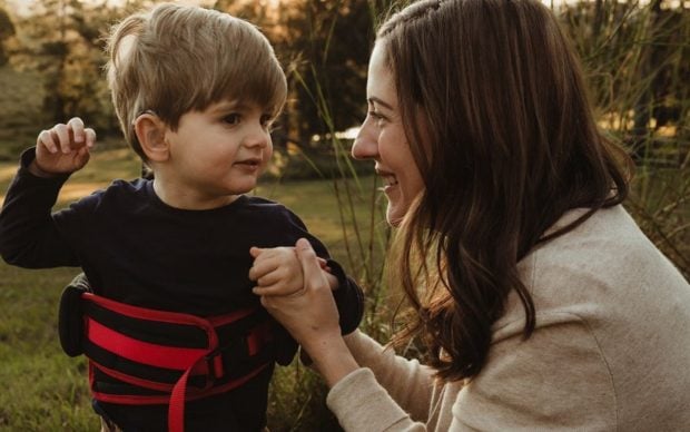 A woman kneels outdoors, smiling and holding hands with a young boy wearing a black shirt and a red support vest. They are surrounded by grass and trees, with sunlight in the background.