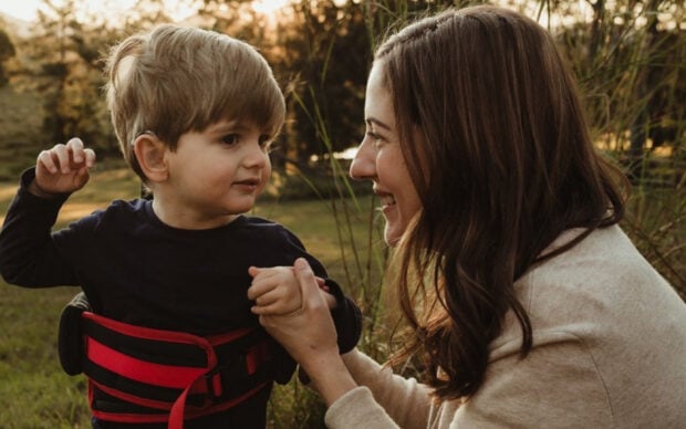 A woman kneels outdoors, smiling and holding hands with a young boy wearing a black shirt and a red support vest. They are surrounded by grass and trees, with sunlight in the background.
