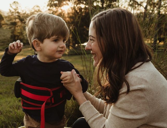 A woman kneels outdoors, smiling and holding hands with a young boy wearing a black shirt and a red support vest. They are surrounded by grass and trees, with sunlight in the background.