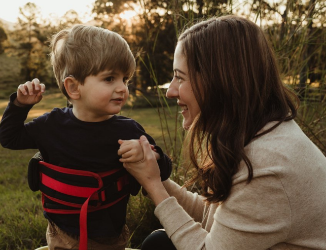 A woman kneels outdoors, smiling and holding hands with a young boy wearing a black shirt and a red support vest. They are surrounded by grass and trees, with sunlight in the background.